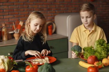 Positive little girl and boy sitting at desk with various fresh vegetables and fruits while preparing meal in loft kitchen with brick wall